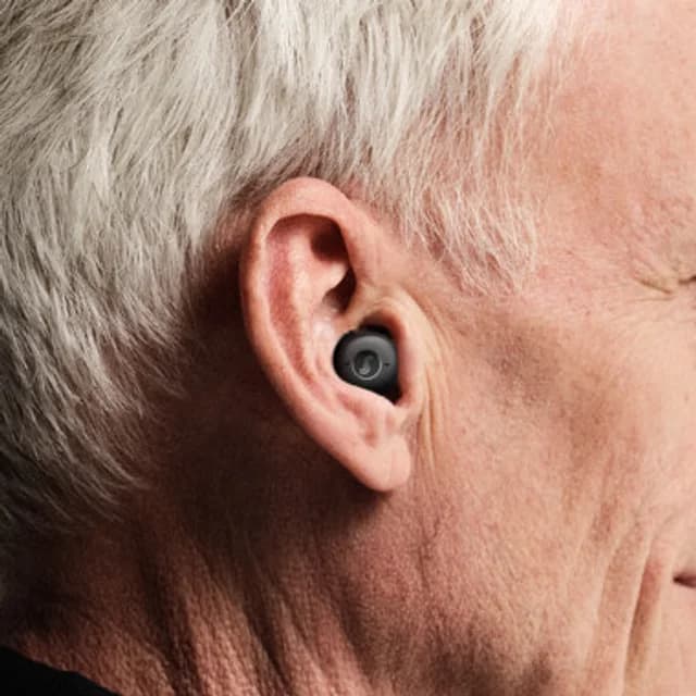 An older man wearing a black earbud-style in-the-ear hearing aid seated in the bowl of his ear