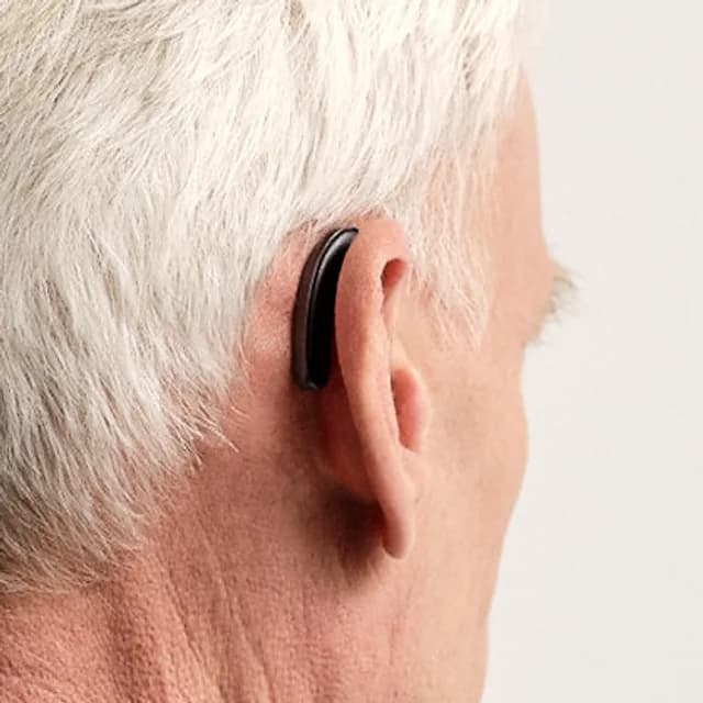 A man with white hair seen from behind, a slim black receiver-in-canal hearing aid tucked discreetly behind his ear